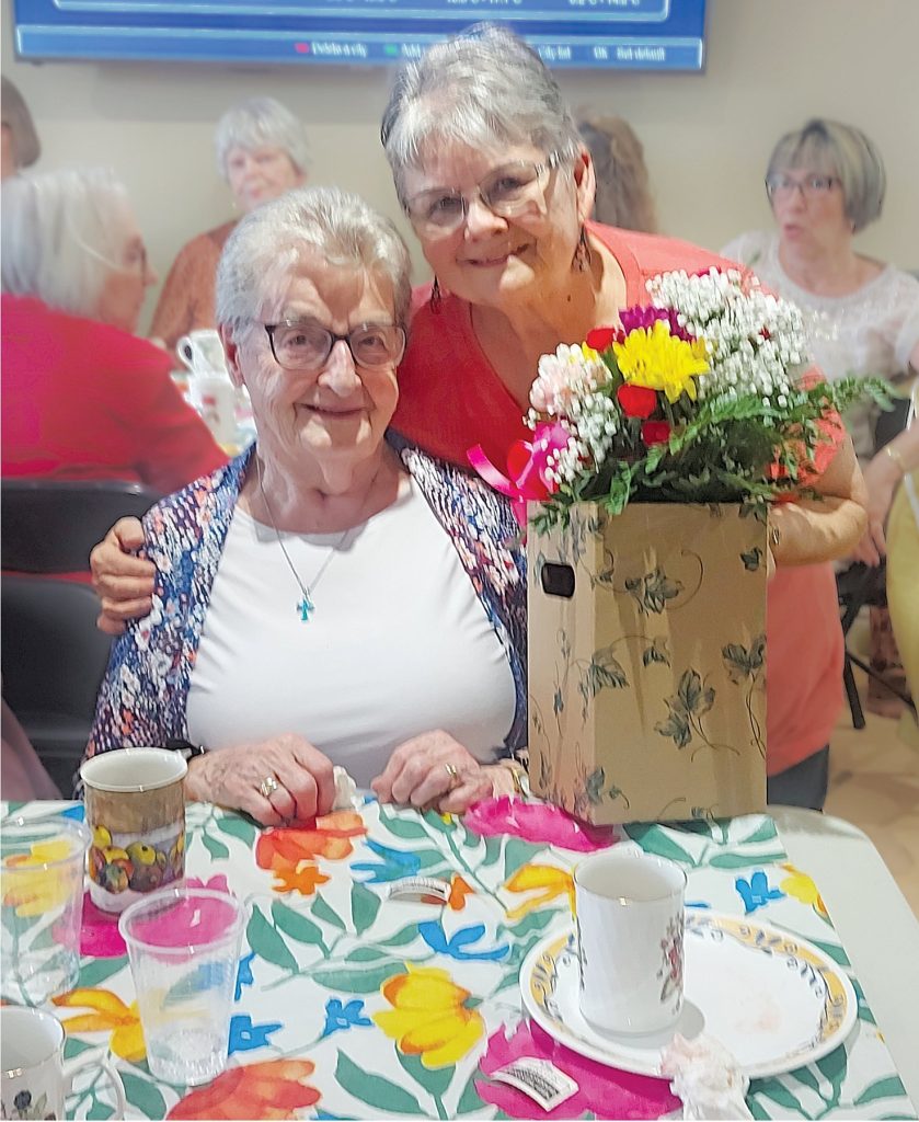 two women with grey hair smile at the camera. One is seated, and the one standing behind her is presenting her with flowers
