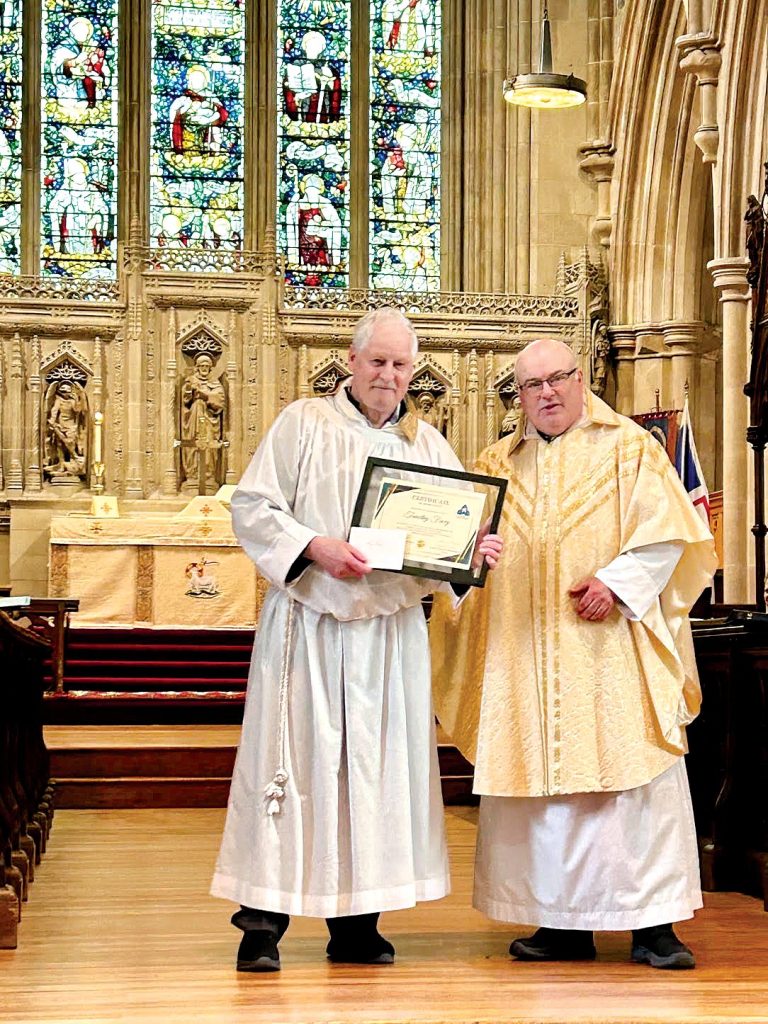Two men stand at the front of a stone church, one dressed as a server and the other as a priest. The one dressed as a server holds a certificate.