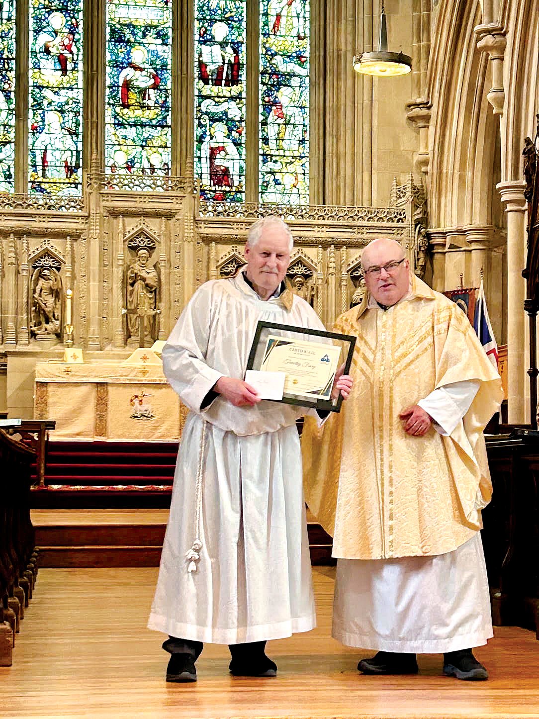 Two men stand at the front of a stone church, one dressed as a server and the other as a priest. The one dressed as a server holds a certificate.