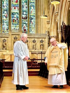 Two men stand at the front of a stone church, one dressed as a server and the other as a priest. The one dressed as a priest is holding a wooden board on which the names a sacristans are written.