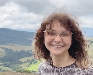 a girl with glasses and shoulder-length wavy hair smiles in front of mountains in the background