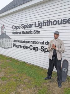A man dressed in dark pants and a light top stands next to a skateboard at the lighthouse in Cape Spear Newfoundland