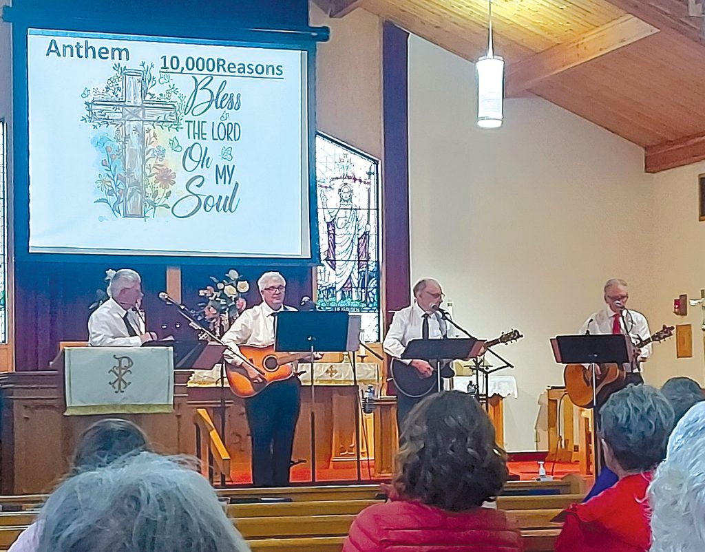 Four men playing acoustic guitars on a church stage below a projection screen.