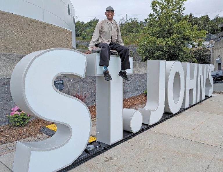 A man dressed in dark pants and a light top sit on top of the "St. John's" sign
