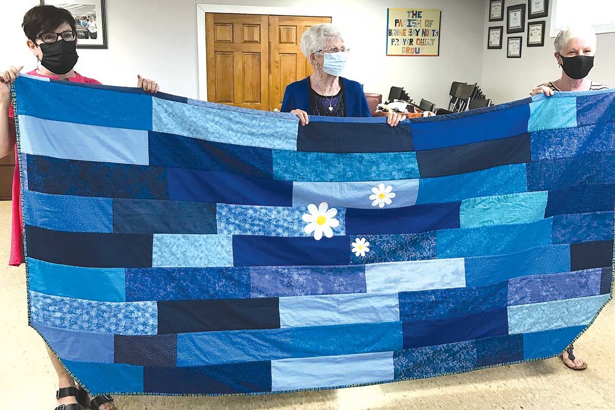 One side of the Daisy/Dignity quilt completed in blues with daisies appliquéd and stitched around perimeter with a fine herringbone stitch (completed by Doris Pittman). Left to right: Gail Kennedy-Sparkes, Doris Pittman, and Linda Parsons