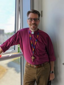 photograph of Bishop Sam Rose wearing rainbow cord cross and leaning on a window